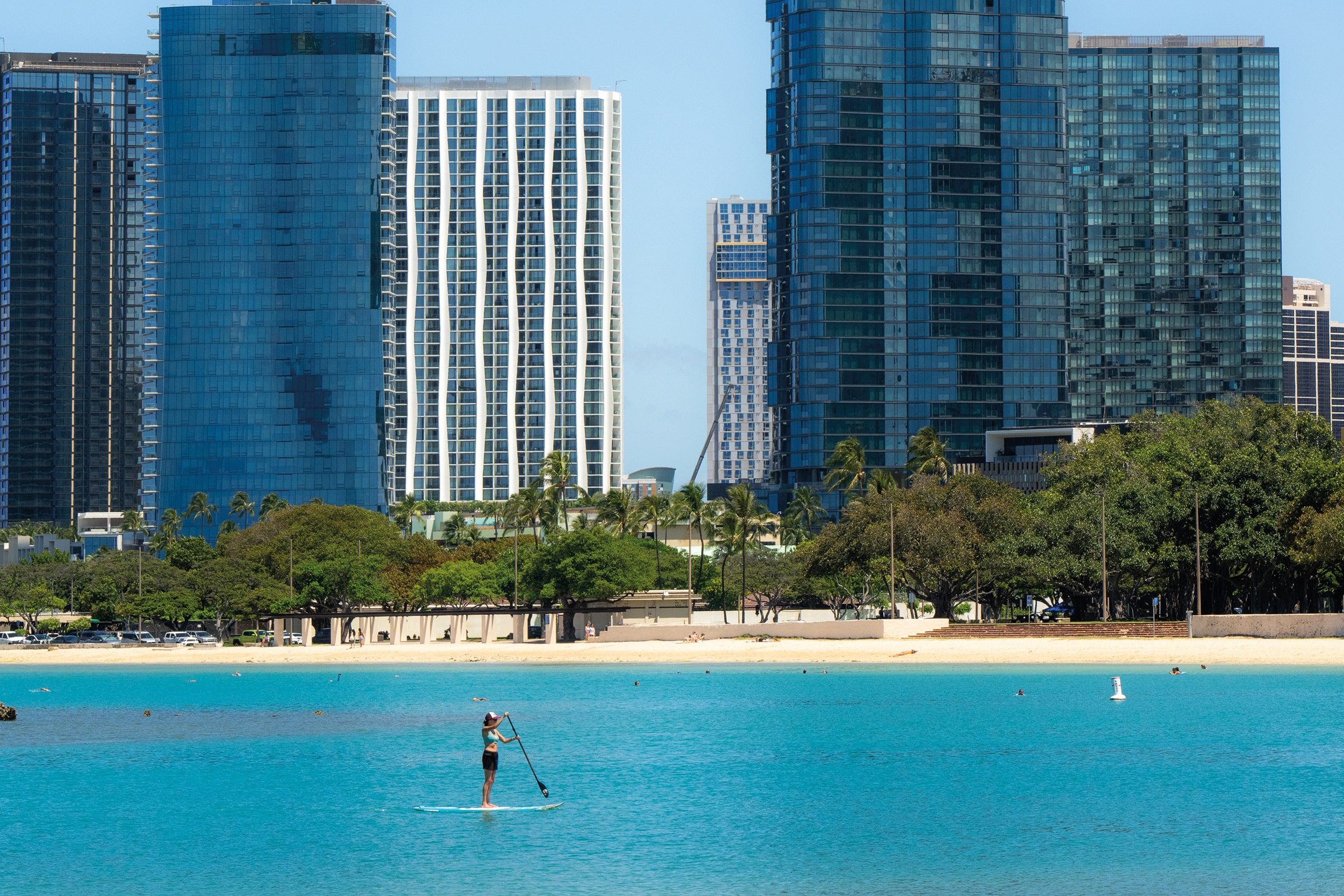 A person paddleboards on turquoise water near a sandy beach, with tall modern buildings and trees in the background, showcasing a blend of urban living and sustainability.