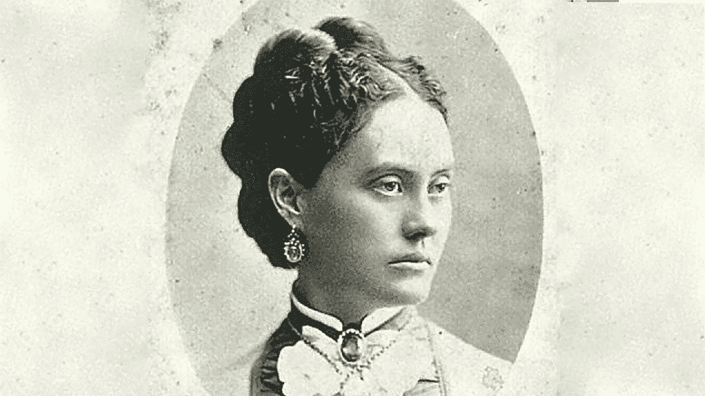 Black and white portrait of a woman with styled hair, wearing earrings, a high-collared dress, and a brooch, looking slightly to the side.