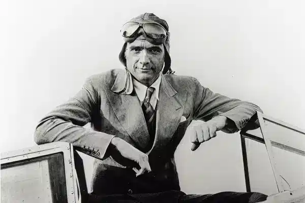 A man in a suit and tie sits on an aircraft, wearing aviator goggles and a leather flight cap, looking directly at the camera.
