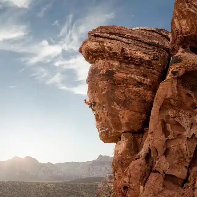 A rock climber ascends a steep, rugged cliff face with a clear sky and distant mountains in the background.