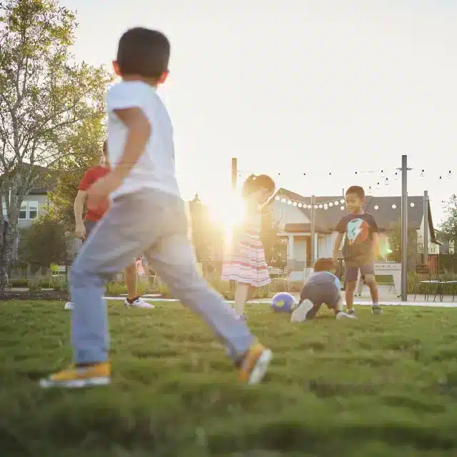 Children play with a ball on a grassy field in a neighborhood park at sunset, with houses and string lights visible in the background.