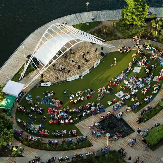 Aerial view of an outdoor amphitheater by the water with people seated on the grass watching a live performance on stage.