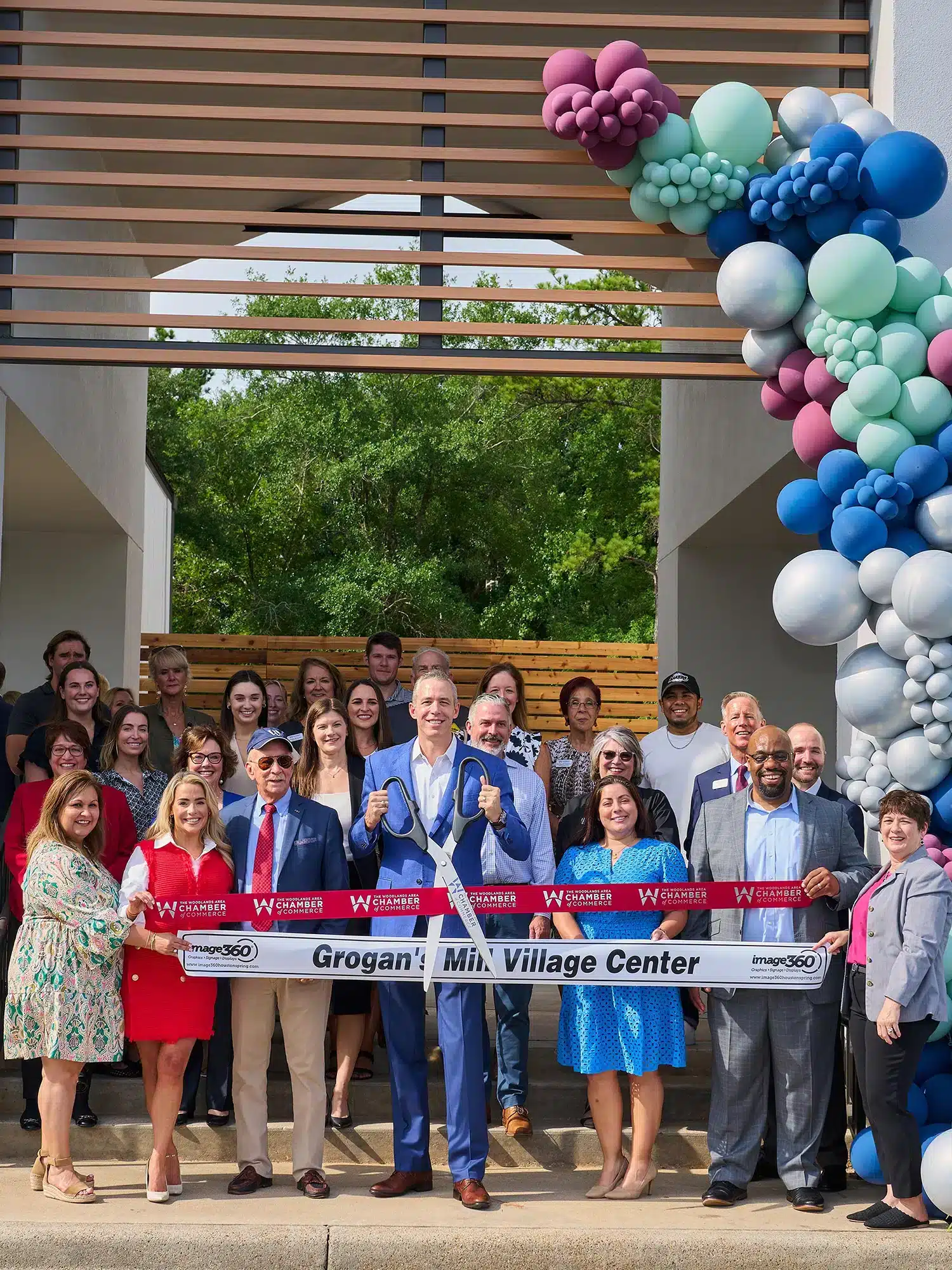 A group of people pose for a ribbon-cutting ceremony in front of a building decorated with balloons, holding a sign that reads "Grogan's Mill Village Center," celebrating its feature in the latest Communities Report.