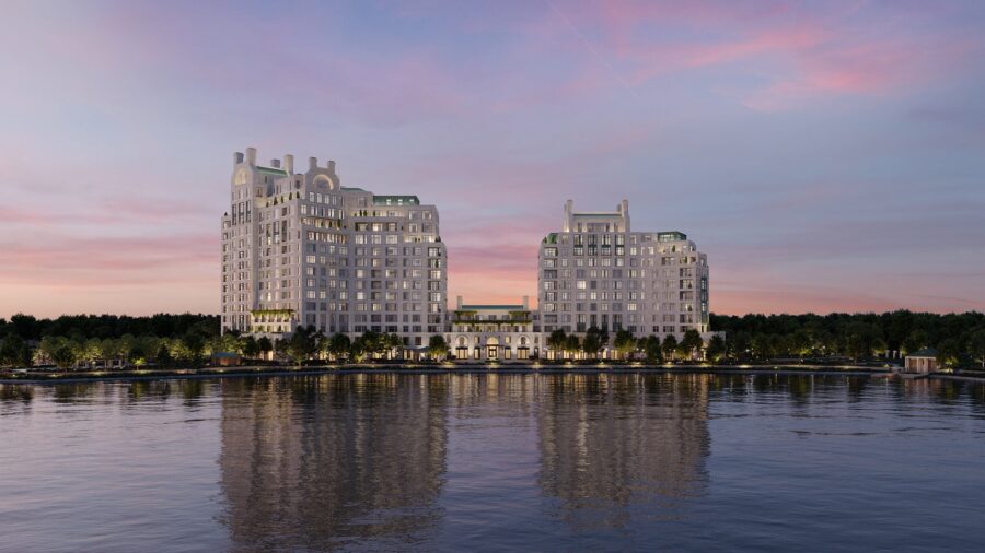 Two symmetrical, mid-rise buildings—part of The Woodlands by Howard Hughes—with green rooftops are reflected in a calm river at sunset, surrounded by trees and a landscaped waterfront.