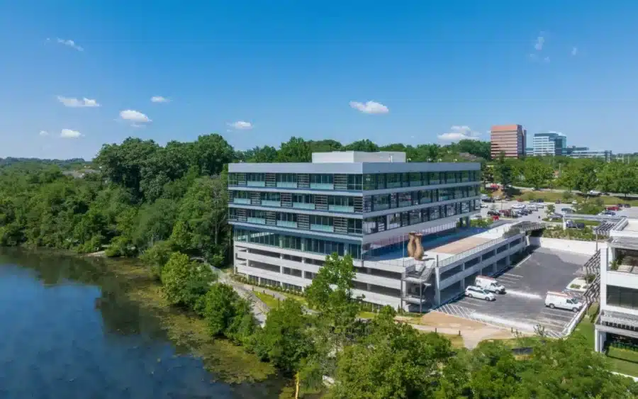 A modern multi-story office building with large windows is situated next to a body of water, surrounded by trees and adjacent parking areas under a clear blue sky.