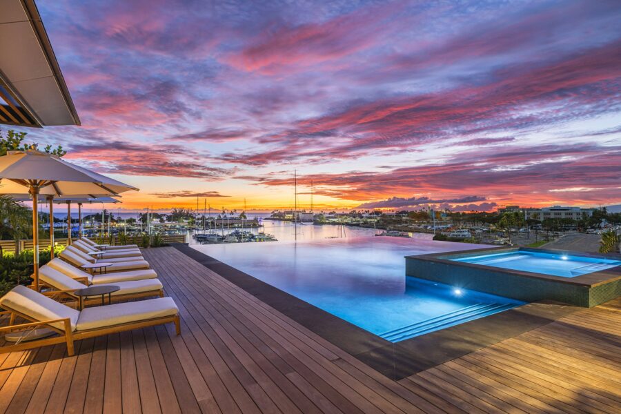 Infinity pool with lounge chairs overlooks the marina at sunset in Ward Village, with boats docked and colorful clouds painting the sky.