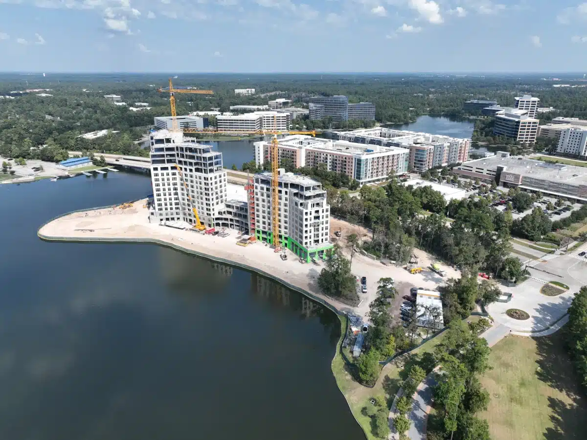 Aerial view of waterfront construction at The Ritz-Carlton Residences, The Woodlands, with several cranes and partially built high-rise buildings near a lake and surrounding Howard Hughes Communities office complexes.