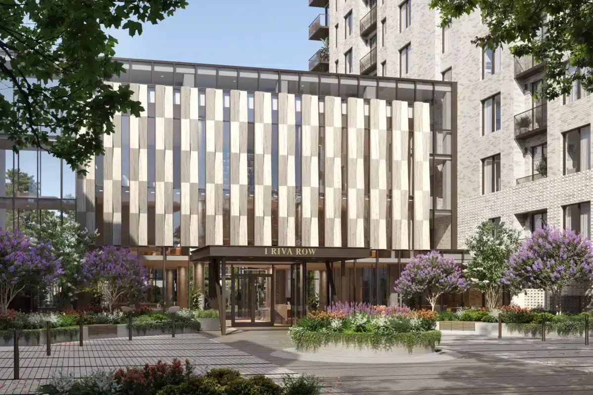 Modern apartment building entrance with vertical wood panels, glass facade, and landscaped planters with purple flowering trees and greenery. Sign above entrance reads 