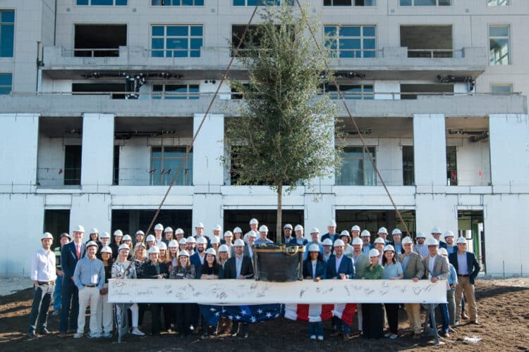 A large group of people in hard hats stands in front of a building under construction at Bridgeland Central, posing with a signed banner and a tree suspended by ropes.