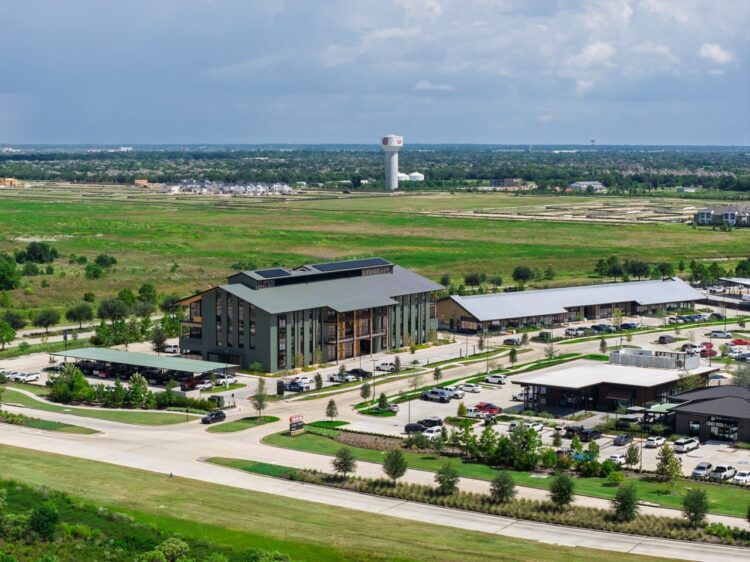 Aerial view of Bridgeland Central business complex with multiple buildings, parking lots, and surrounding green fields under a partly cloudy sky. Retail Arrivals and a distant water tower are visible on the horizon.
