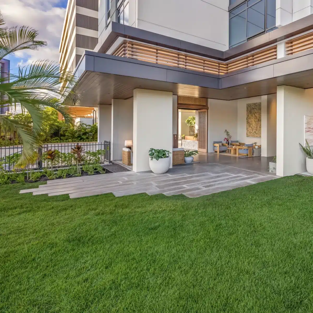 Modern building with a covered patio, potted plants, and large glass doors opening onto a neatly manicured lawn and landscaped garden.