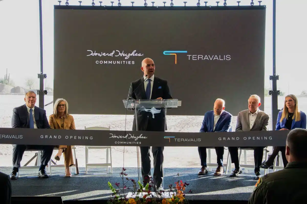 David O'Reilly speaks at a podium during a grand opening event in Phoenix West Valley, with five people seated behind him and a screen displaying Howard Hughes Communities and Teravalis logos.