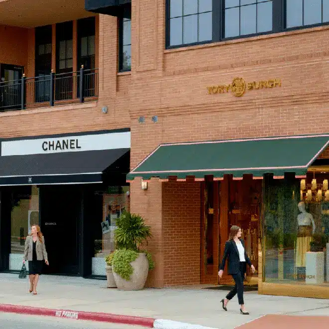 Two women walk past the storefronts of Chanel and Tory Burch, located side by side in a shopping area with brick and beige walls.