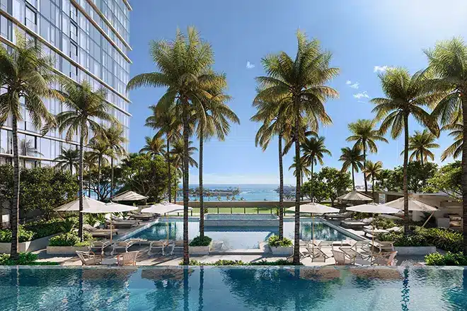Outdoor swimming pools surrounded by palm trees, lounge chairs, and umbrellas, with a modern building on the left and an ocean view in the background under a clear blue sky.