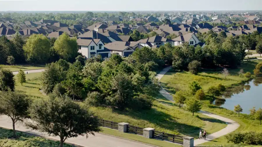 A suburban neighborhood with rows of houses, tree-lined streets, a winding walking path, and a small pond bordered by greenery.