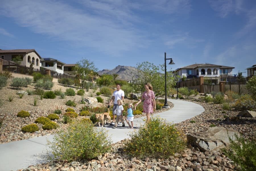 A family with two children and a dog walks along a curved paved path, surrounded by desert landscaping and mountains in the background.