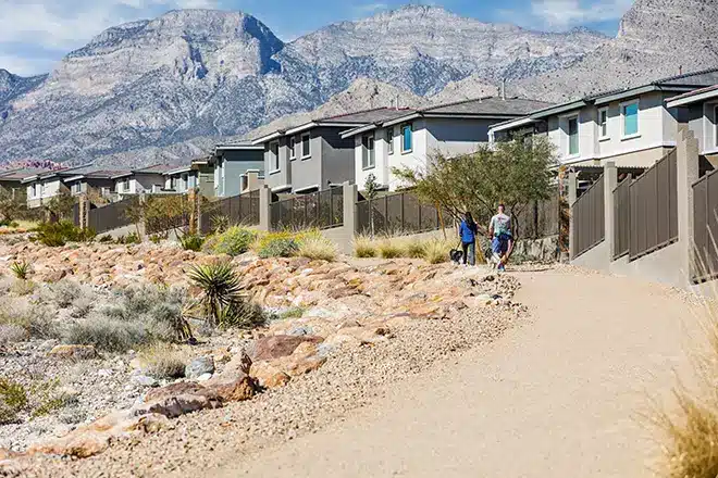 A paved walking path runs alongside a row of modern houses with mountains in the background; two people and a dog walk on the trail.