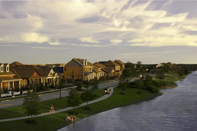 A row of suburban houses lines a winding street beside a lake, with people walking and cycling on a sidewalk under a partly cloudy sky.