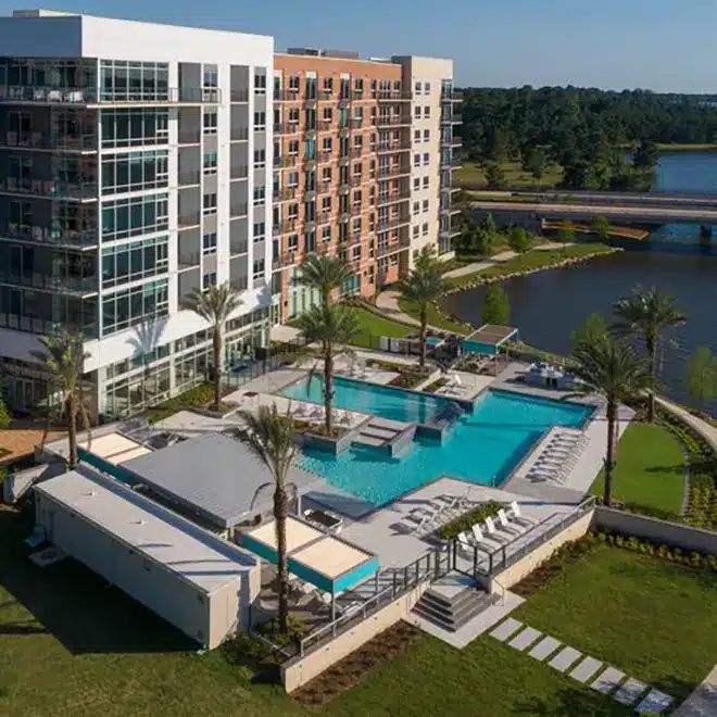 A modern apartment complex with a large outdoor swimming pool, lounge chairs, palm trees, and a view of a nearby lake and wooded area.