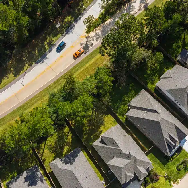 Aerial view of a suburban neighborhood with houses, green lawns, and trees bordered by a road with two cars driving in opposite directions.