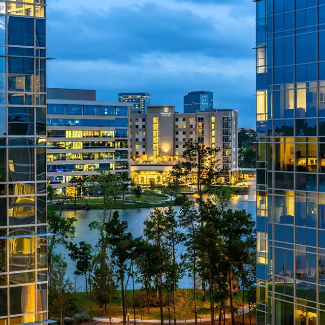 View of modern office buildings and a hotel overlooking a lake, with trees and a walking path in the foreground under a cloudy evening sky.
