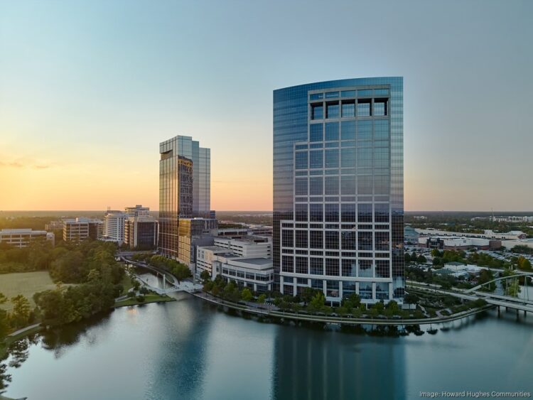 Two modern glass office towers stand beside a calm lake at sunset, surrounded by trees and other buildings in an urban setting.