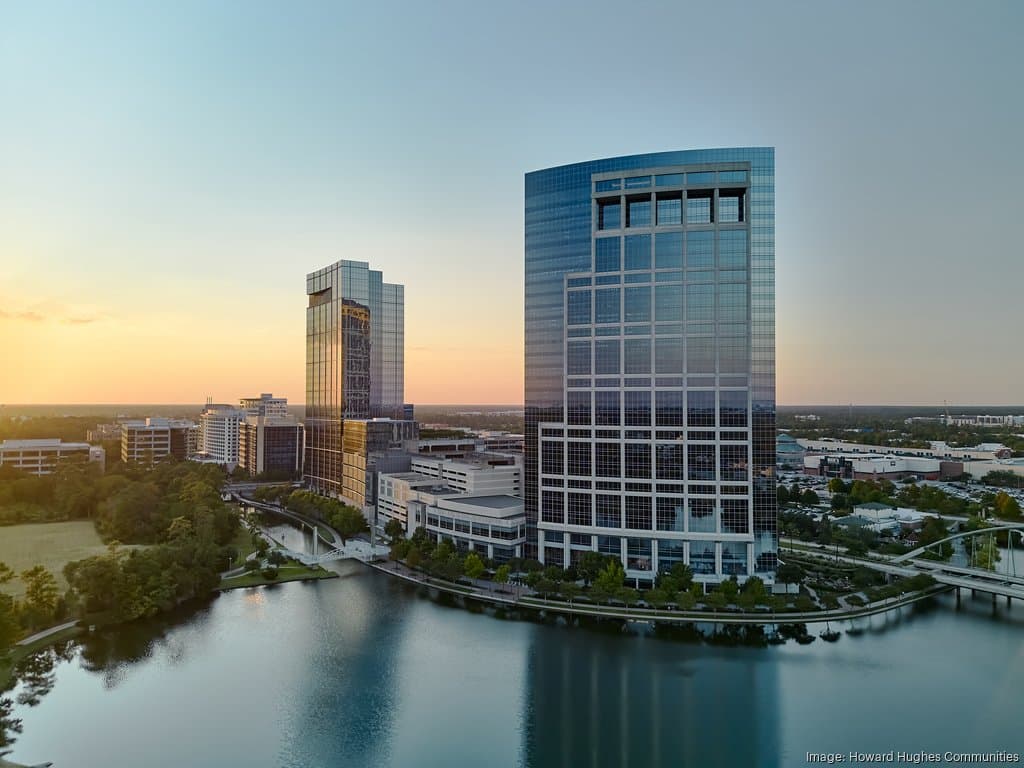 Two modern glass office towers stand beside a calm lake at sunset, surrounded by trees and other buildings in an urban setting.