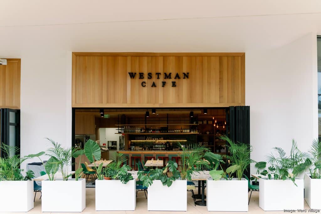 The entrance of Westman Cafe features large windows, potted plants, and neatly arranged tables and chairs outside, with a view of the interior counter and seating area.