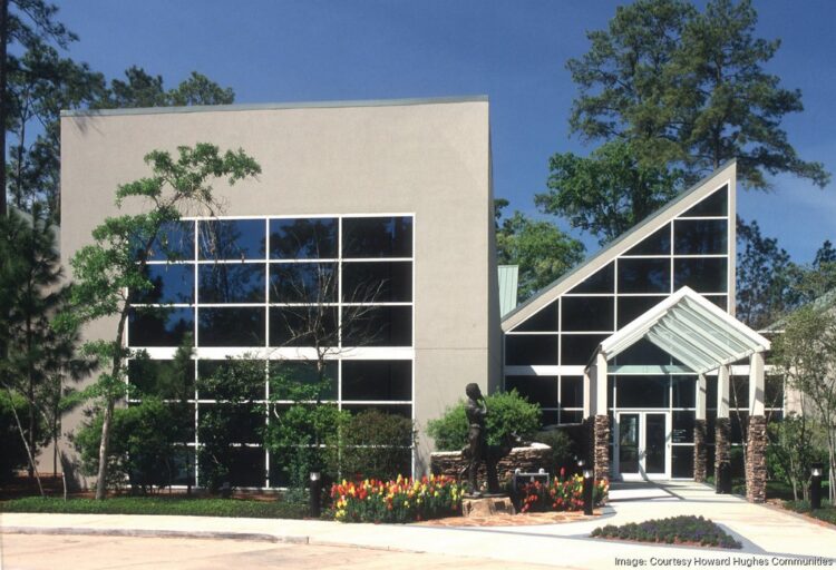 Modern office building with large glass windows, geometric design, and landscaped entrance with flowers and trees under a clear sky.