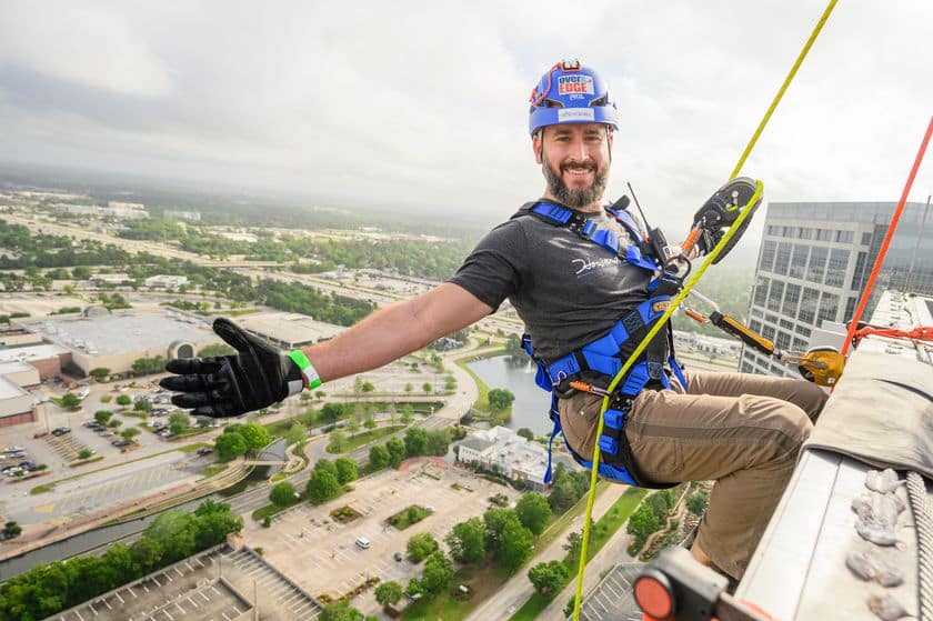 A man wearing safety gear and a helmet rappels down the side of a tall building, smiling and spreading his arms, with a cityscape visible in the background.