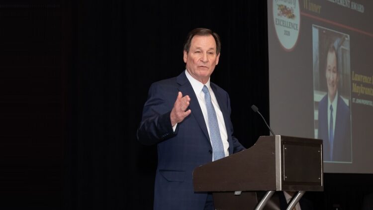 A man in a suit speaks at a podium with a microphone; an award graphic and a photo are projected on a screen behind him.