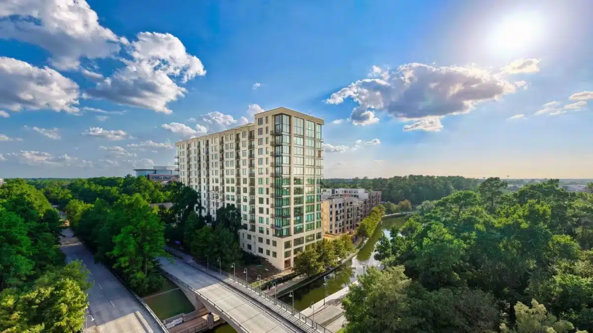 A tall modern apartment building stands near a small waterway, surrounded by trees and roads, under a bright blue sky with scattered clouds.