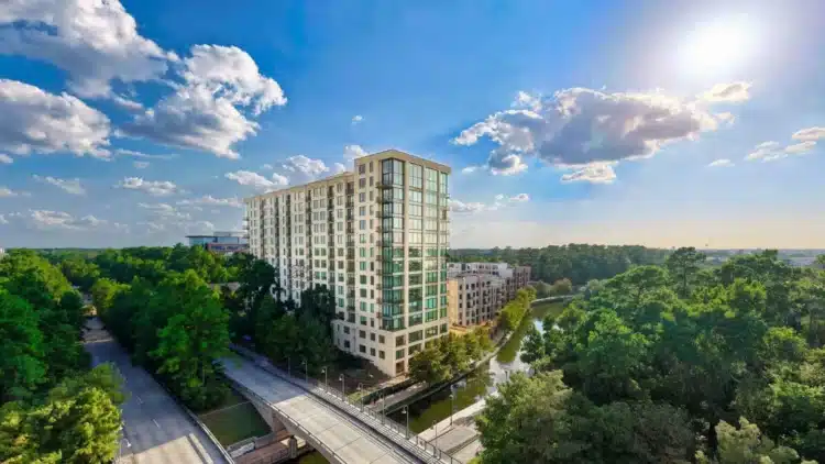 A tall modern apartment building stands near a small waterway, surrounded by trees and roads, under a bright blue sky with scattered clouds.