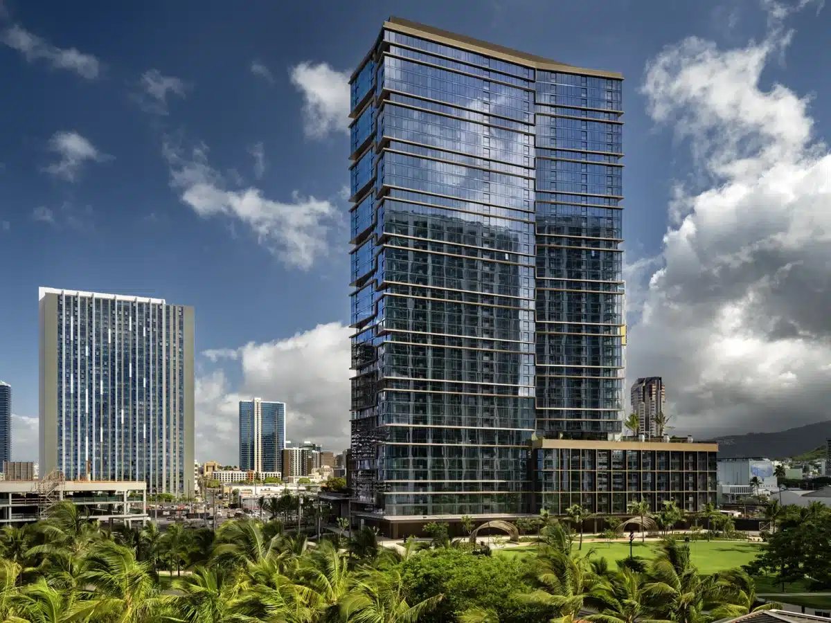 A modern high-rise building with glass windows stands among other tall buildings, set against a partly cloudy sky and palm trees in the foreground.
