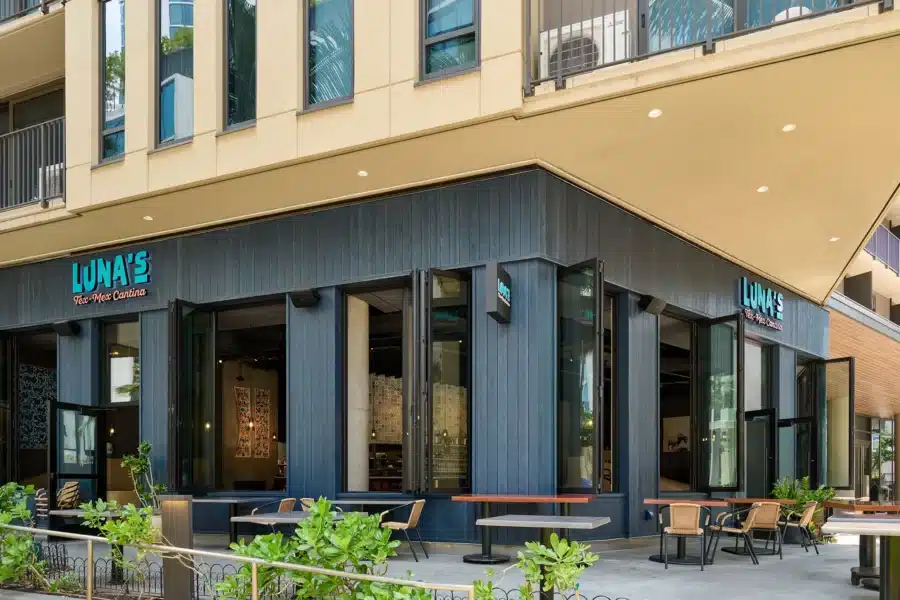 Corner view of Luna’s restaurant with large windows, outdoor seating, blue signage, and modern architectural design. Tables and potted plants are arranged near the entrance.