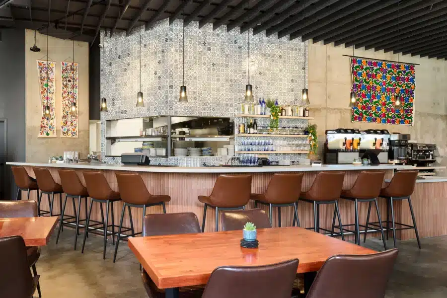 Modern restaurant interior with a tiled bar, brown chairs, wooden tables, and colorful wall art. Pendant lights hang from the ceiling, and drink machines are visible on the right.