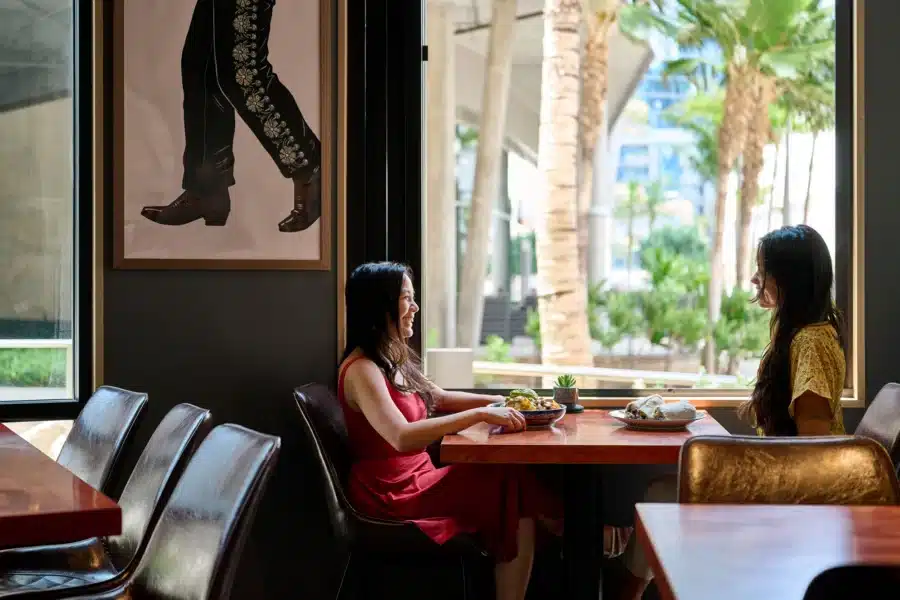 Two women sit across from each other at a restaurant table with food. Large windows show palm trees outside. A framed photo of legs in boots hangs on the wall.