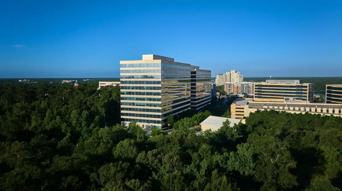 A glass office building stands among trees, with several other modern buildings nearby under a clear blue sky.