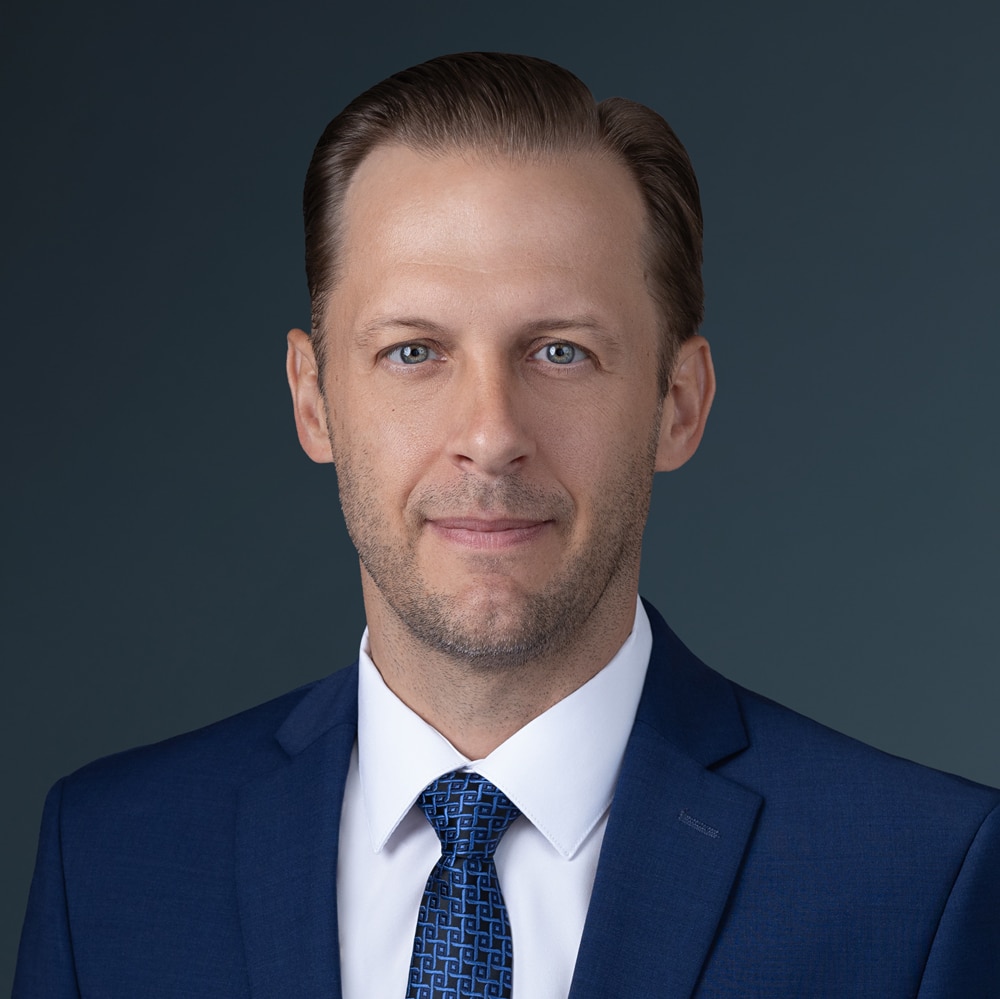 Man in a blue suit and patterned tie, with short brown hair, light complexion, and a neutral expression, posing against a plain dark background.