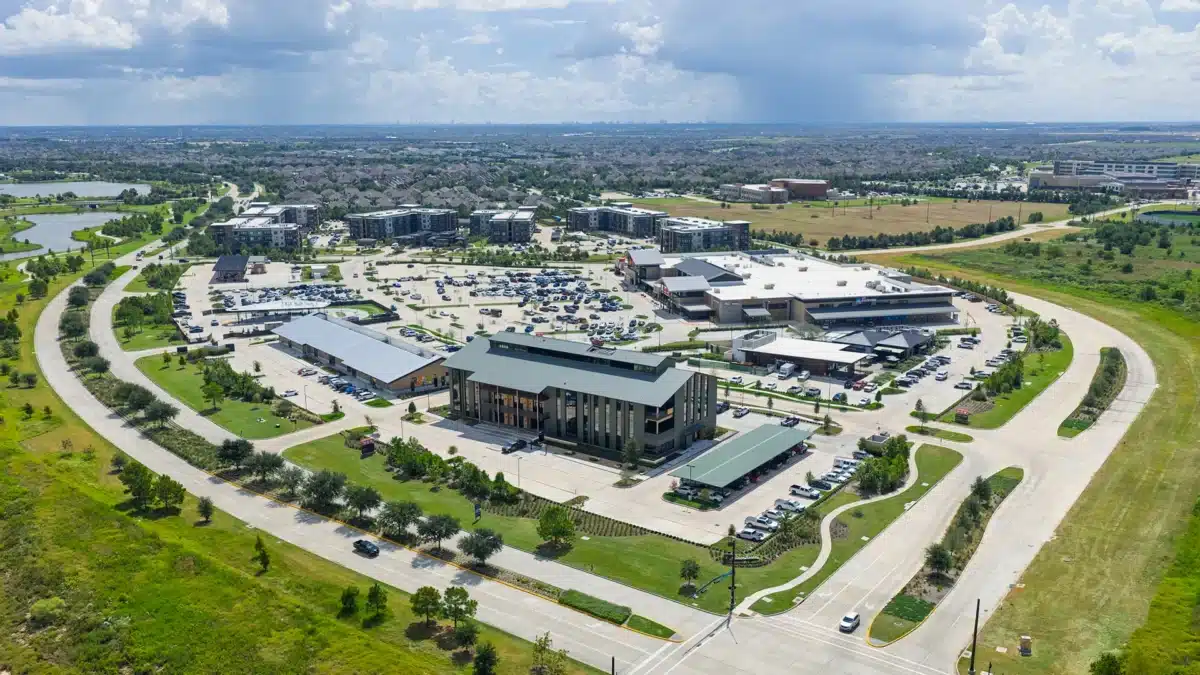 Aerial view of a large commercial complex with multiple buildings, parking lots, and surrounding roads, set in a suburban area with green spaces and distant residential neighborhoods.