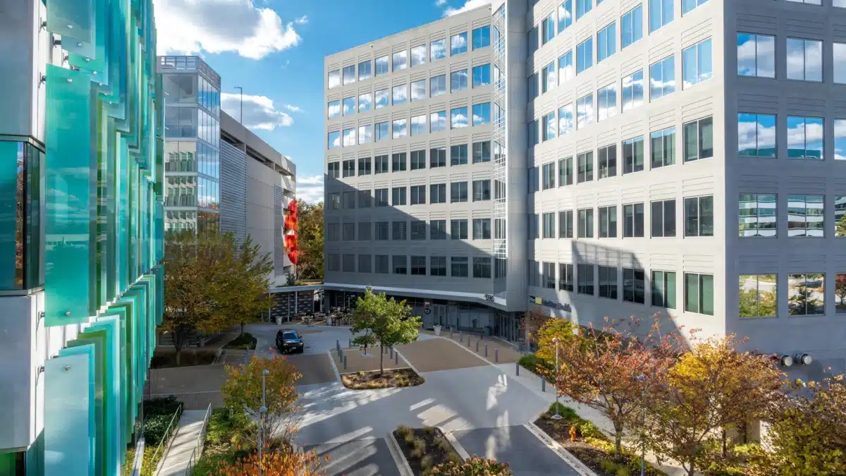 Modern office buildings surround a landscaped courtyard with trees and pathways on a sunny day, under a blue sky with scattered clouds.