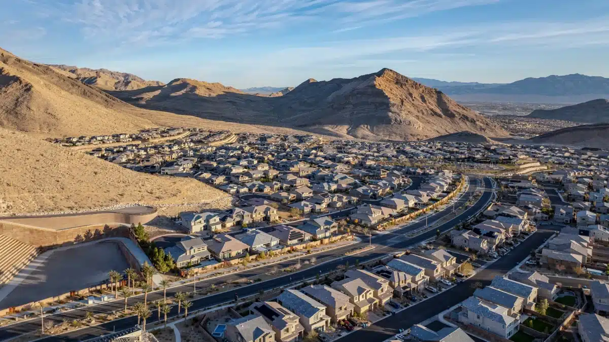 Aerial view of a suburban neighborhood with rows of houses, curving roads, and mountainous desert landscape in the background under a clear sky.