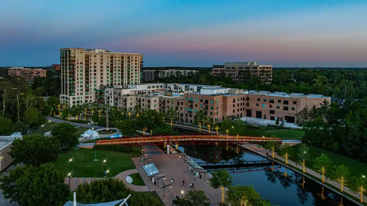 Aerial view of a city park with a pond, red pedestrian bridge, walking paths, green trees, and multi-story residential buildings at dusk.