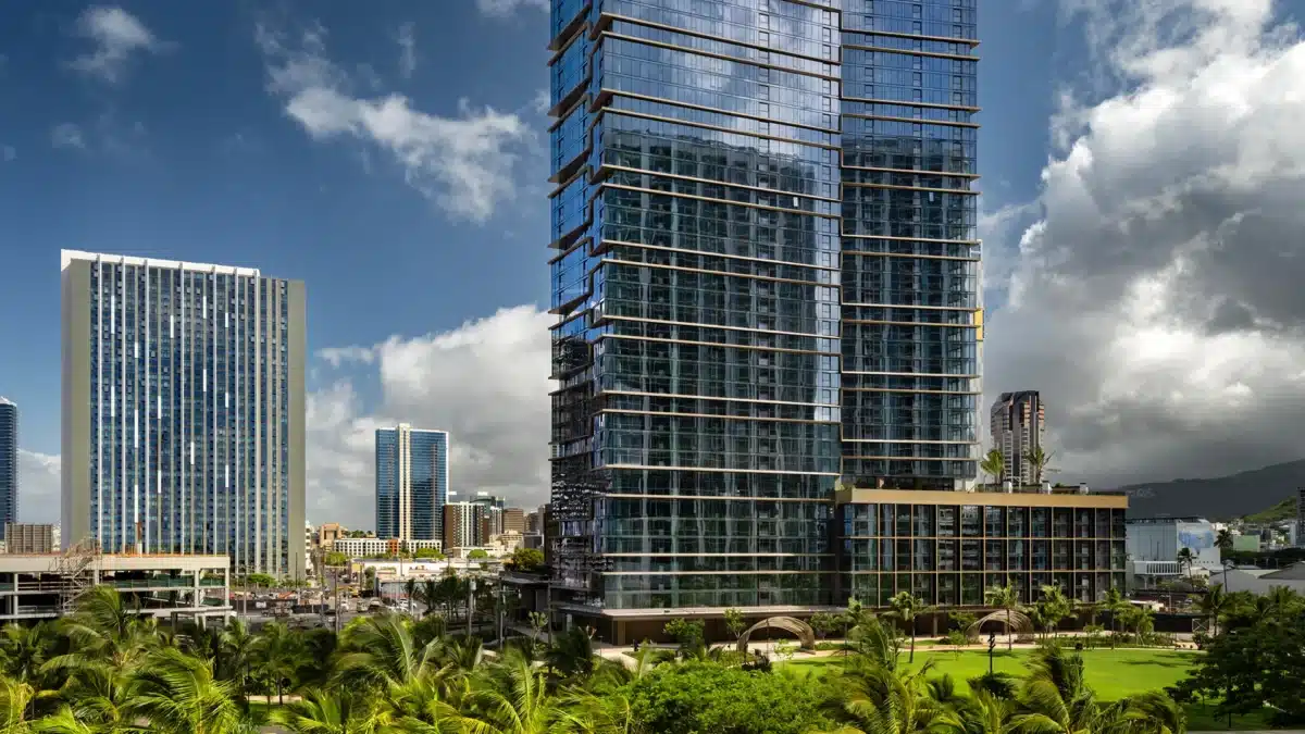 Tall modern glass buildings rise above palm trees and greenery under a partly cloudy sky in an urban cityscape.