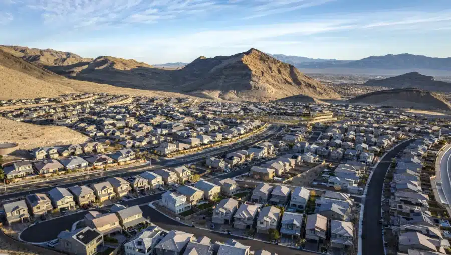 Aerial view of a suburban neighborhood with rows of houses, surrounded by dry hills and mountains under a clear blue sky.