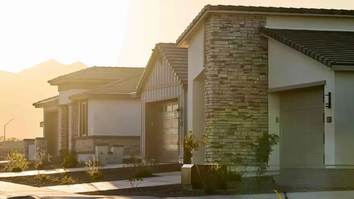 Row of modern single-story houses with stone and stucco exteriors, driveways, and small front yards, photographed at sunset.