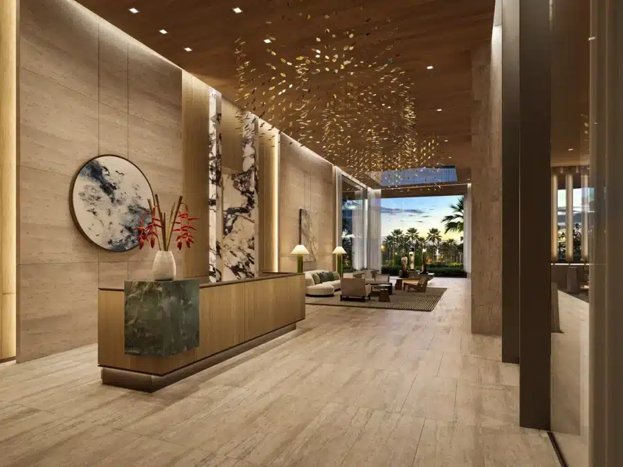 Modern hotel lobby with marble floors, wooden ceiling, contemporary art, reception desk, lounge seating, and large windows showing a view of trees and palm trees outside.