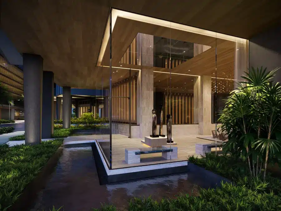 Modern building entrance at night with large glass windows, wooden ceiling, sculptures inside, water feature, and surrounding greenery.