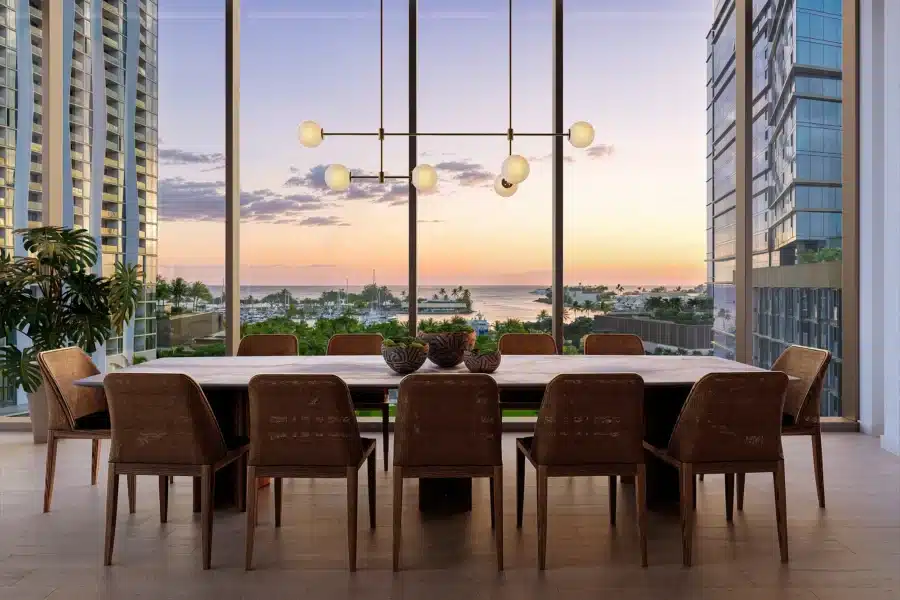 Modern dining room with a long table and ten chairs, large windows showing a sunset view of the ocean, marina, and city buildings. Contemporary chandelier hangs above the table.