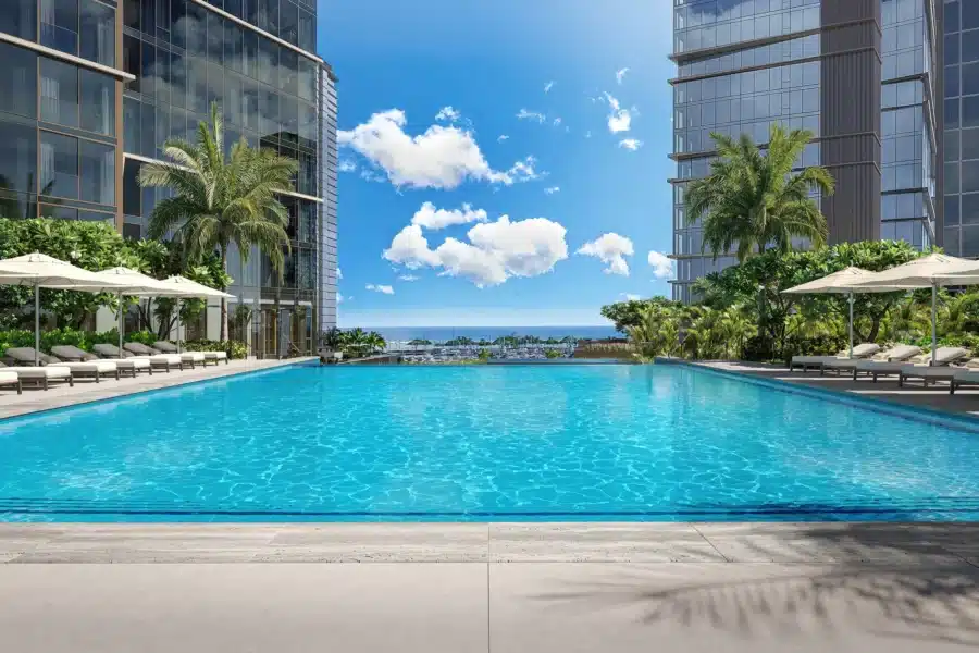Outdoor swimming pool between two modern high-rise buildings, surrounded by palm trees and lounge chairs, with a view of the ocean and a blue sky with clouds.